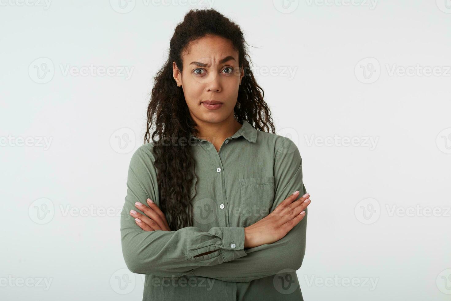 Confused young attractive dark haired curly female keeping her hands crossed and raising surprisedly eyebrow while looking at camera, standing over white background photo