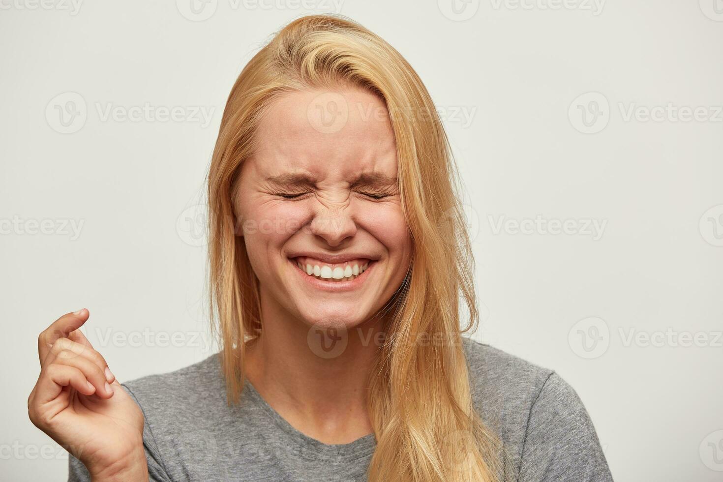 cerca arriba de riendo hermosa rubia joven mujer, siente divertido, Escuchó un broma o gracioso historia, cerrado su ojos, con dientes sonrisa, usa gris camiseta, aislado terminado blanco antecedentes foto