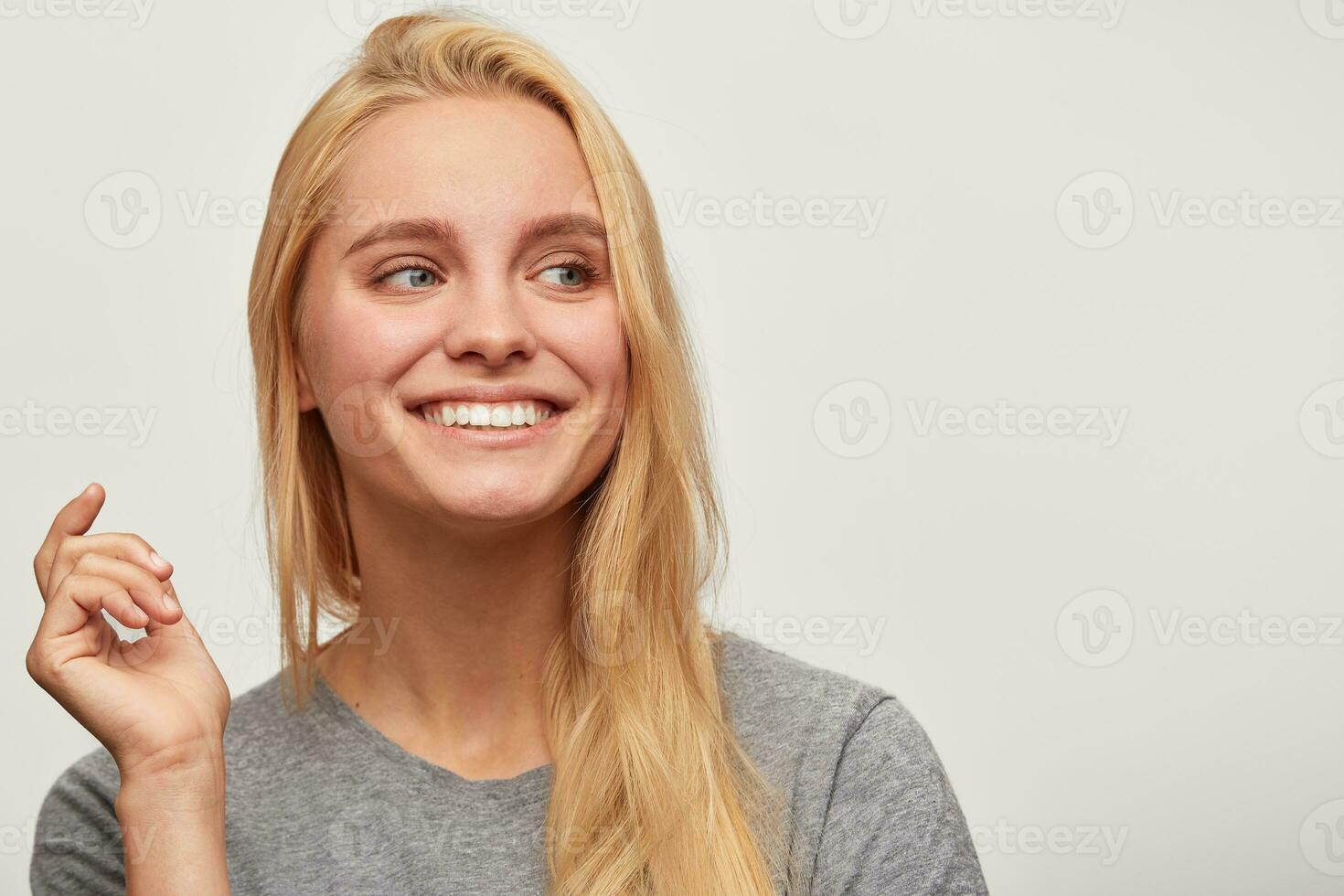 Portrait of laughing nice blonde young woman, spends time with close friends company, feels happy, one hand up, looking aside, toothy smile, wears grey t-shirt, isolated over white background photo