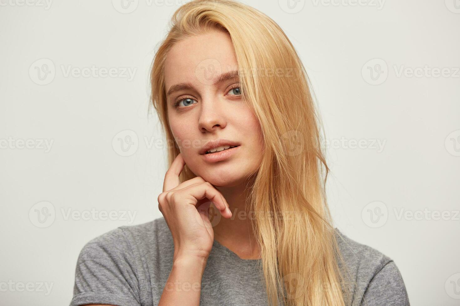 Portrait of lovely beautiful mysterious blonde young woman with blue eyes holds her hand near chin, attentively listens and looks at the interlocutor isolated over white studio background looks camera photo