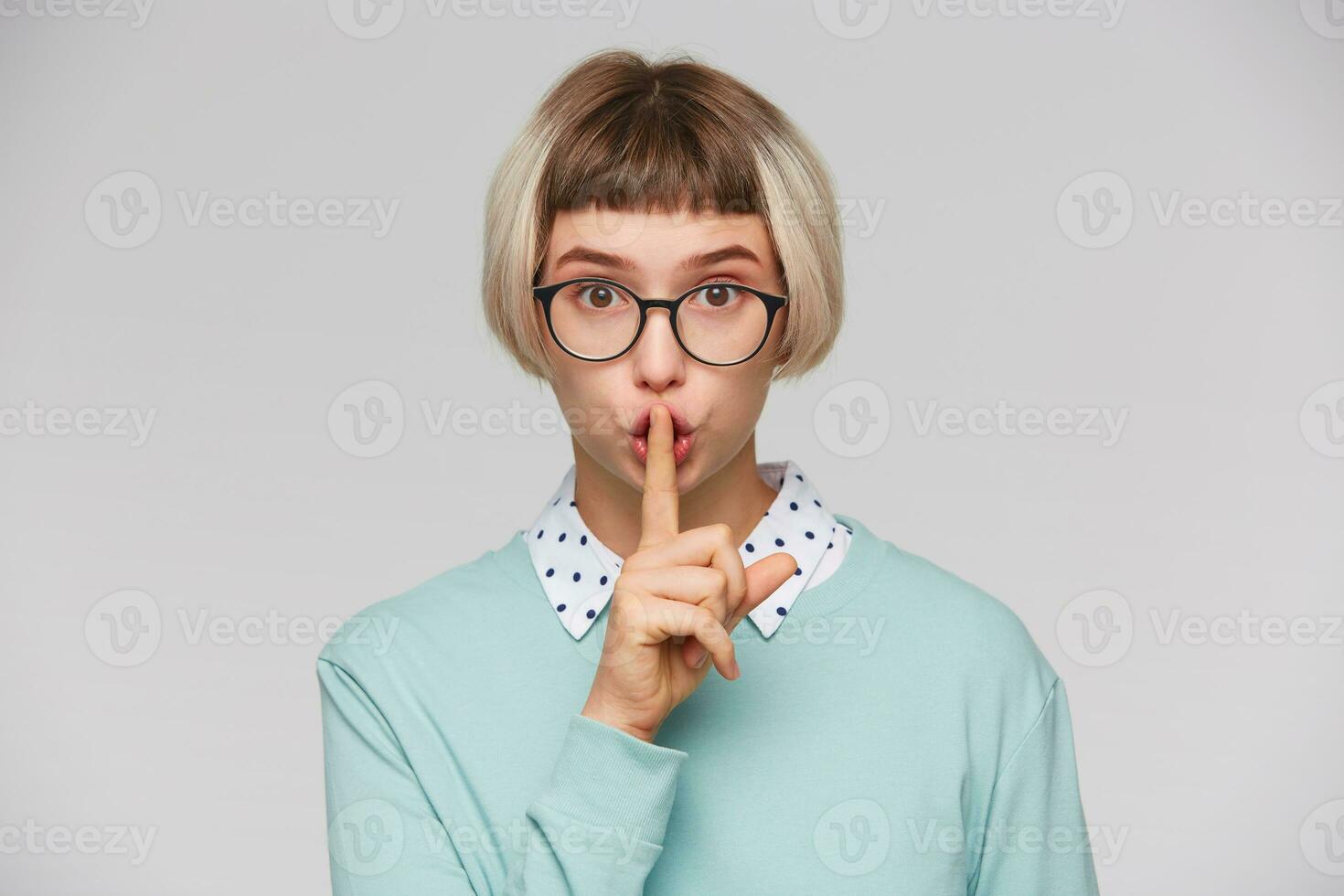 Closeup of cute beautiful young woman wears blue sweatshirt and glasses looks concentrated and shows silence sign isolated over white background photo