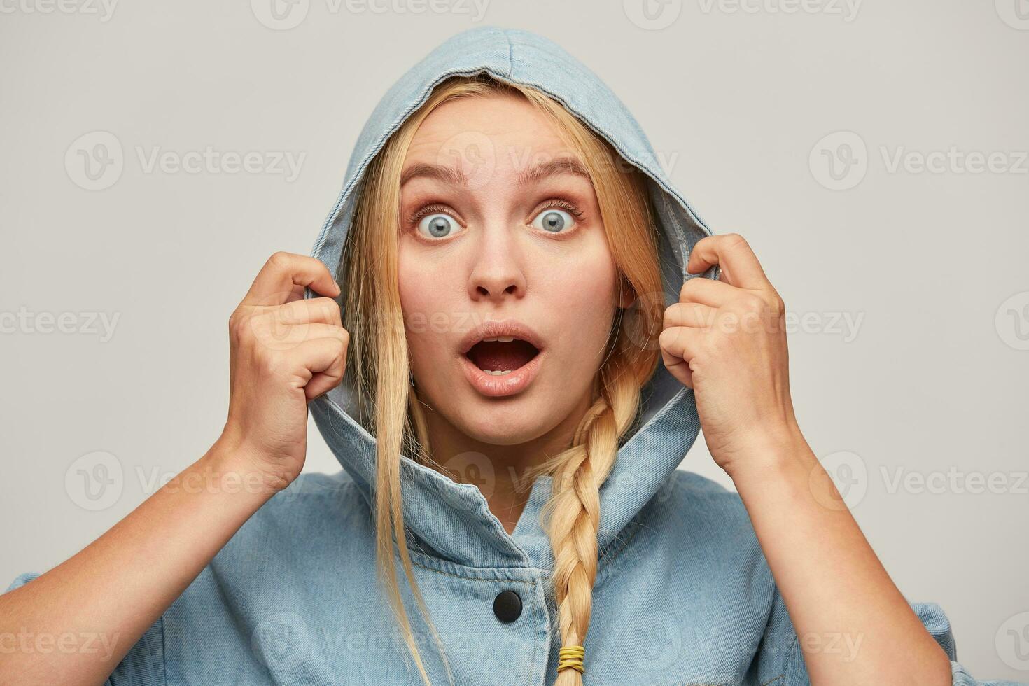 Portrait of amazed beautiful blonde young woman with braid, hands keep hood, looks open-eyed, wears oversize denim coat very surprised or shocked, over white background photo