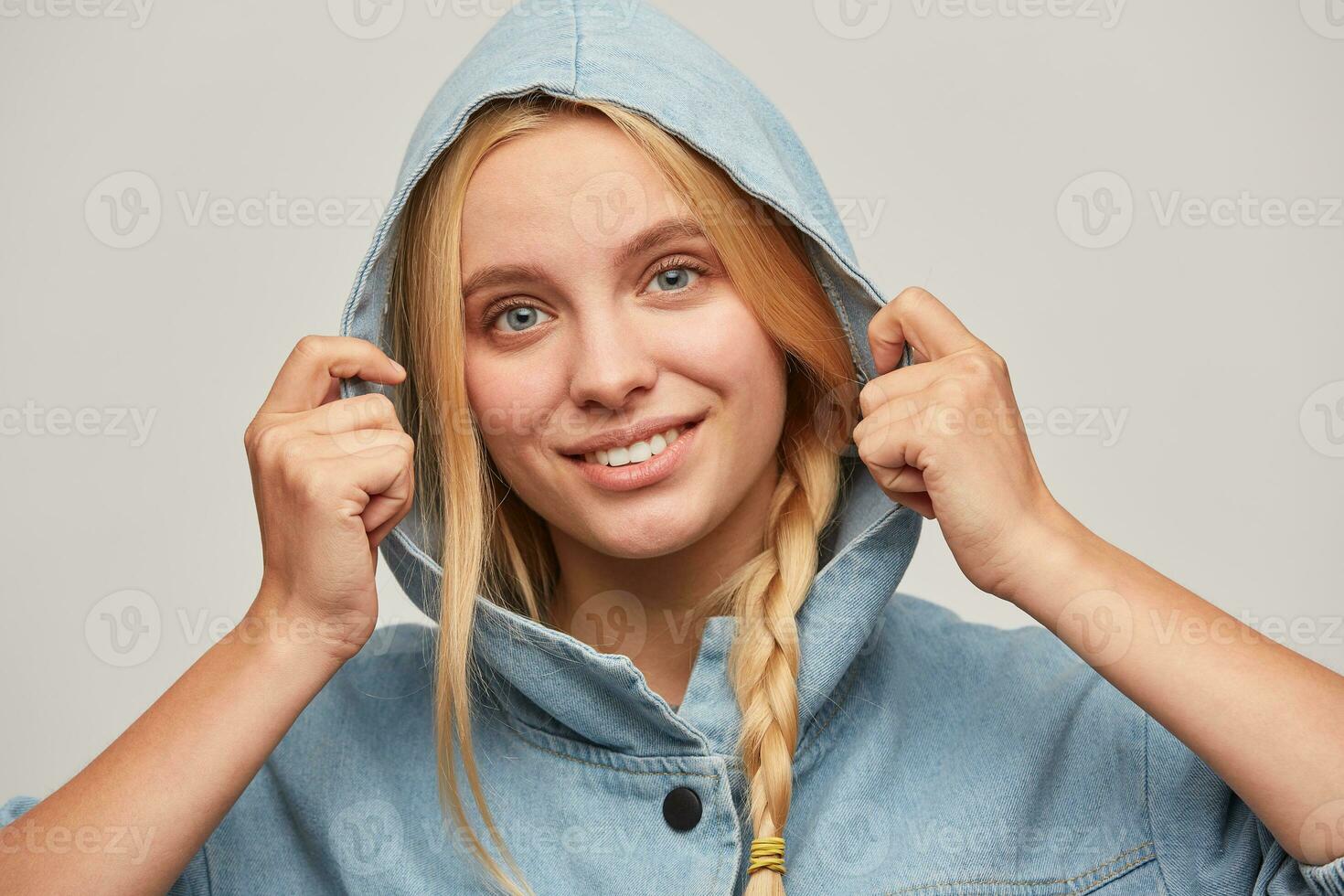 Tender beautiful blonde young woman with braid, hands keep hood, feels happy, smiles to a viewer, wears oversize denim coat feeling warmed, going to a pleasant meeting, over white background photo