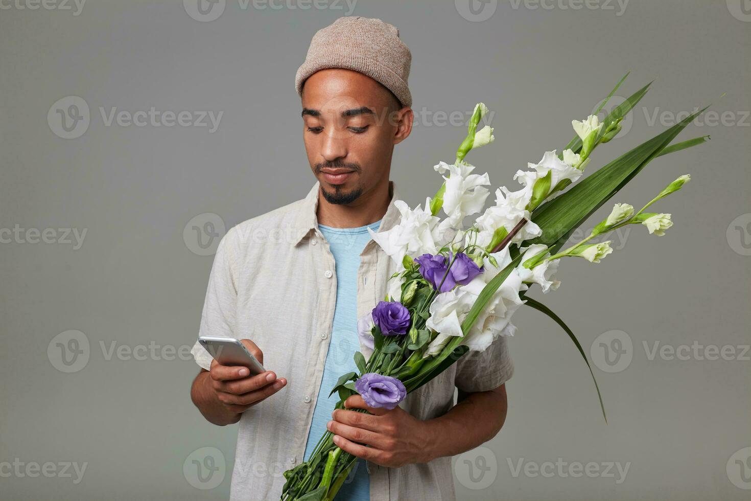 Portrait of young cheerful attractive guy in gray hat, holds a bouquet in his hands, looks at the smartphone and chatting, stands over gray backgroud. photo