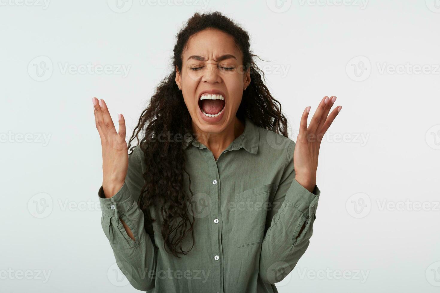 Stressed young curly brunette dark skinned woman raising emotionally her hands while screaming with closed eyes, standing over white background in casual wear photo