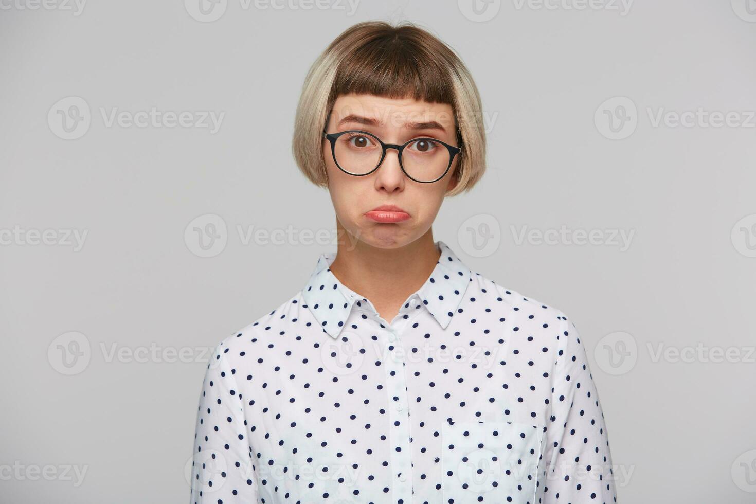 Closeup of sad upset blonde young woman wears polka dot shirt and spectacles curving lips and feels depressed isolated over white background photo