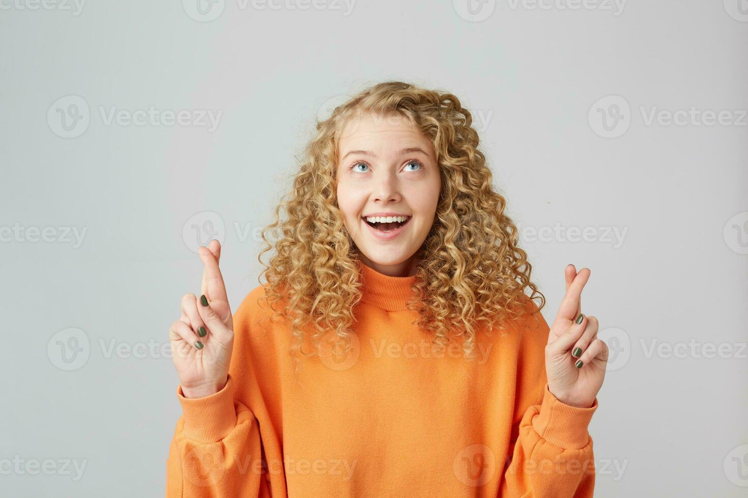 encantador positivo niña atentamente creyendo en su suerte. Disparo de joven Rizado contento sonriente inspirado rubia hembra acuerdo su dedos cruzado y ojos mirando arriba mientras haciendo un deseo foto