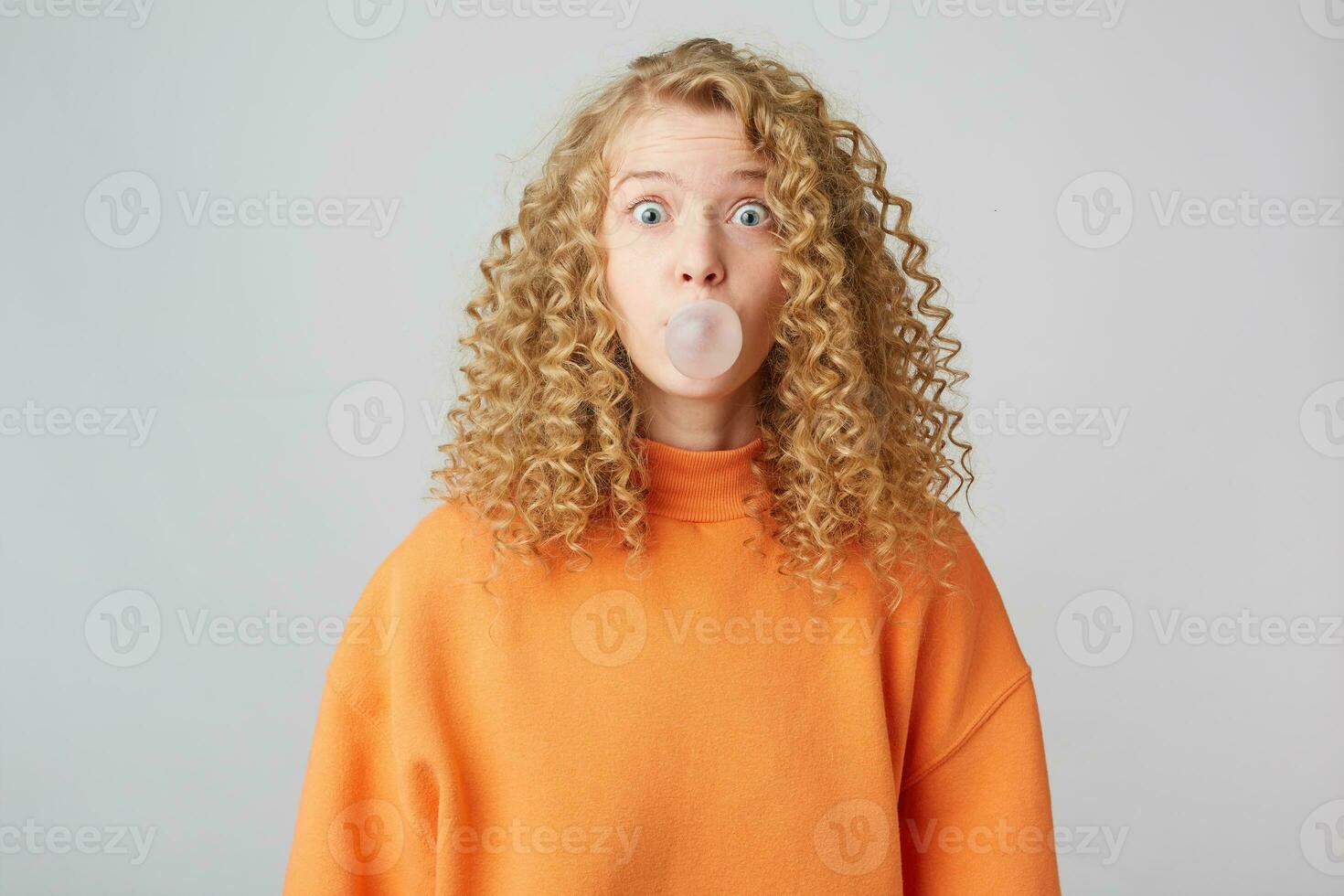 Misunderstanding, naive curly blonde lady dressed in warm orange oversize sweater looking camera isolated over white background while blowing bubble with chewing gum photo