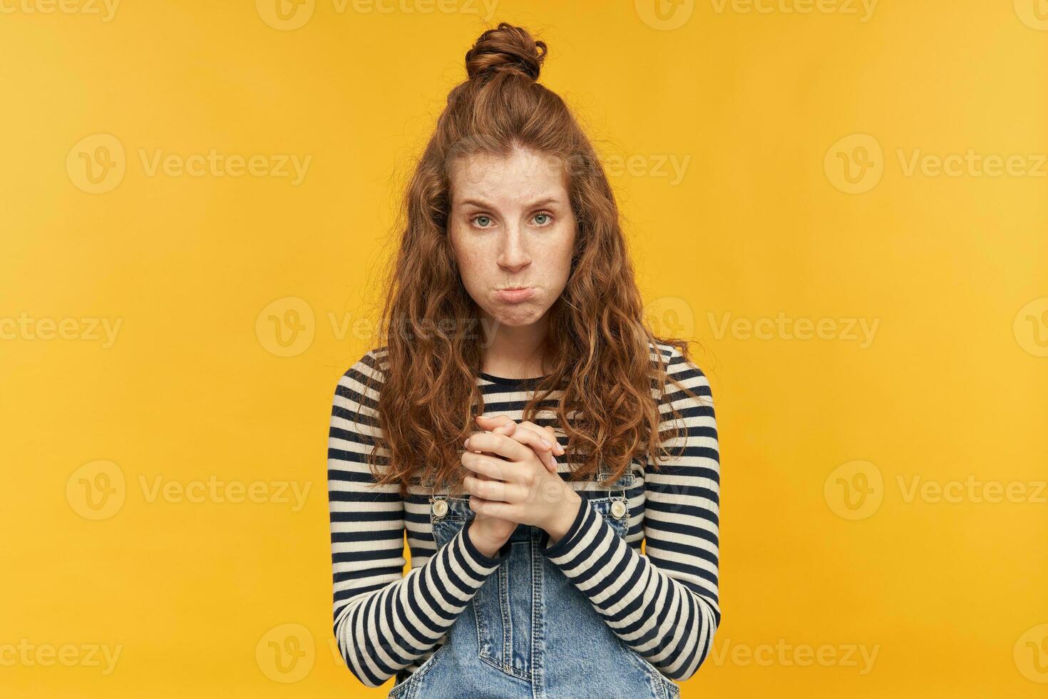 indoor portrait of young displeased ginger girls, keeps her palms together, curves her lips with a sad facial expression. isolated over yellow background. photo