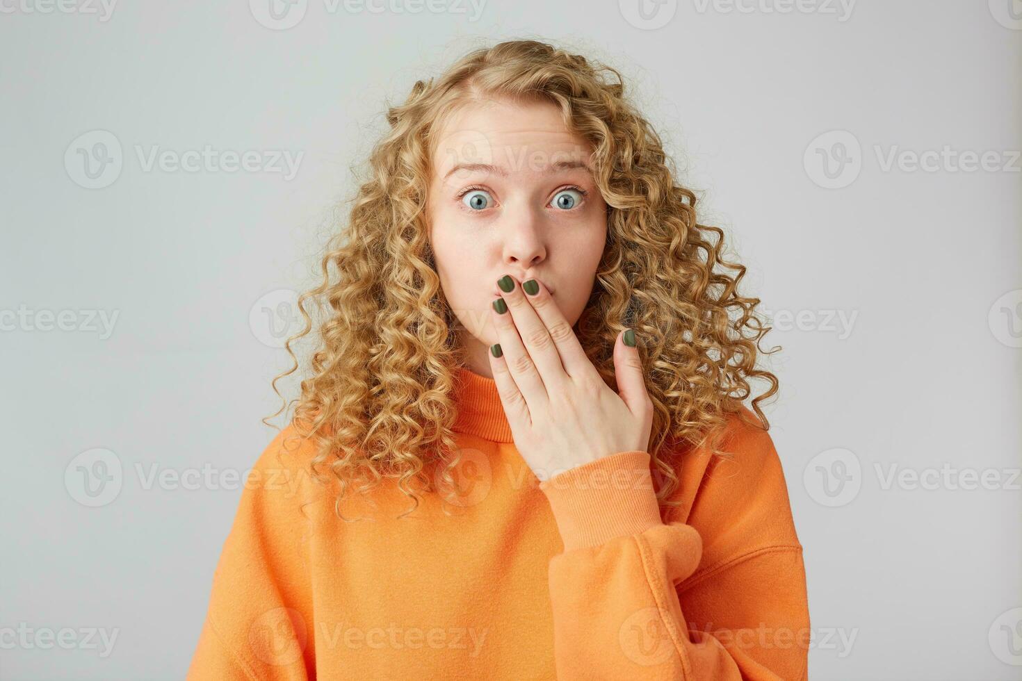 pretty and attractive curly-haired blonde stands staring at the camera, covers her mouth with her hand, as if saying something extra, inappropriate, has revealed someone's secret. photo