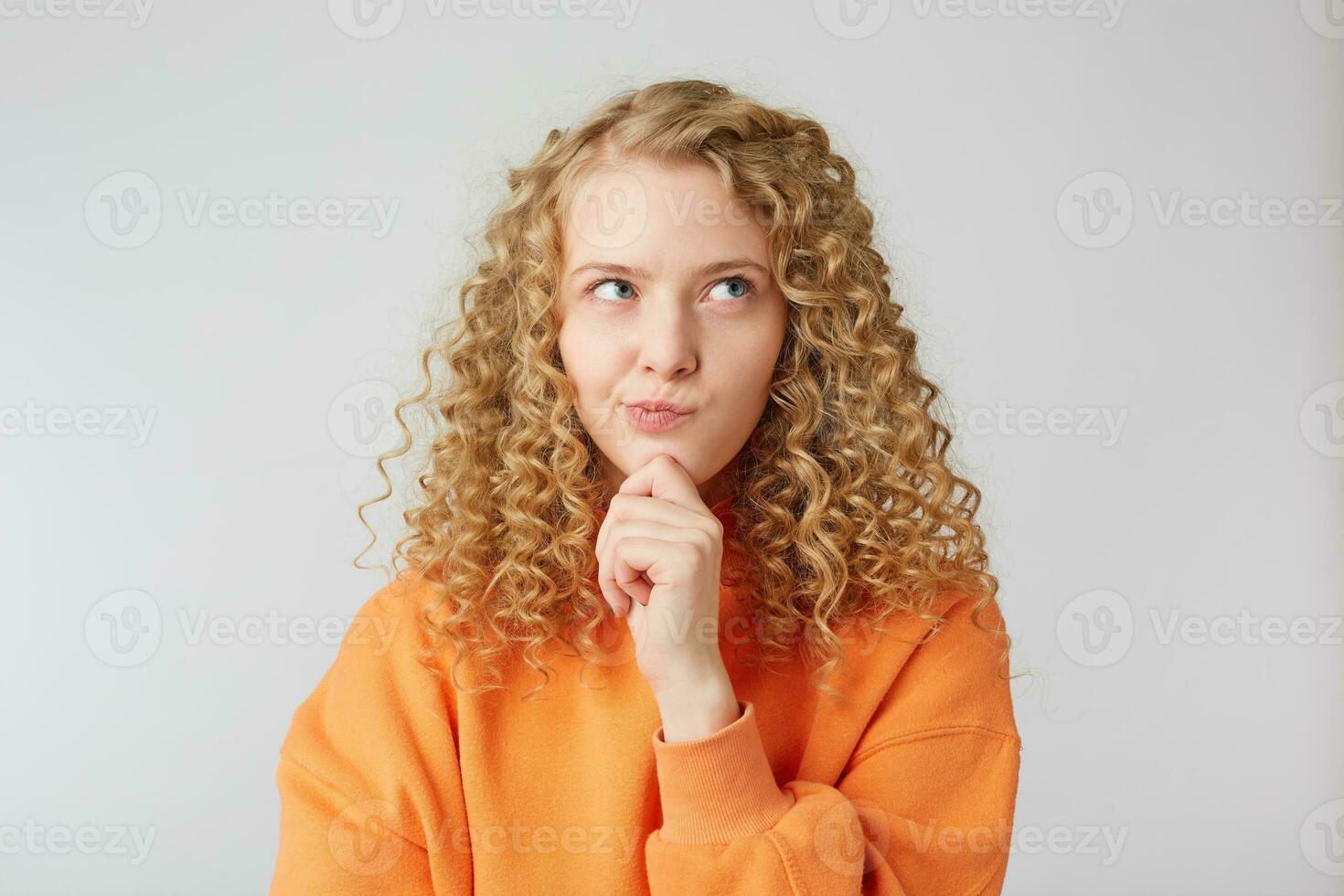 Pondering thoughtful blonde thinking about something, makes machination, looks rightside, makes plans,purses her lips, holds her fist near her chin, wears orange sweater isolated over white background photo