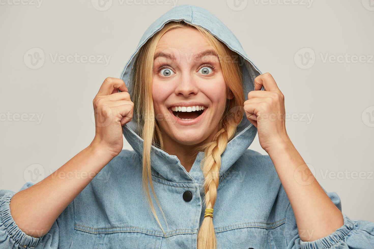 Clouse up of glad beautiful blonde young woman with braid, hands keep hood, smiles, wears oversize jeans denim coat feeling warmed and happy because of good weather, over white background photo