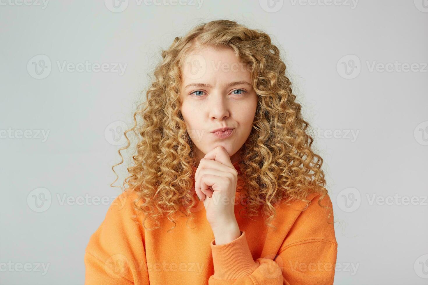 Thoughtful blonde looking into the camera with narrowed eyes, slyly, makes plans,purses her lips, holds her fist near her chin, wears orange sweater isolated over white background photo