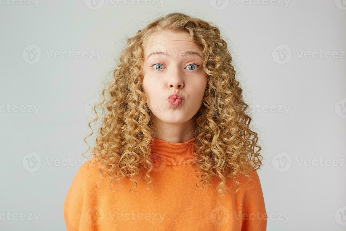 Heartful sweeet lovely curly blonde sends air kiss to a carmera, dressed in warm orange oversize sweater, isolated over white studio background photo