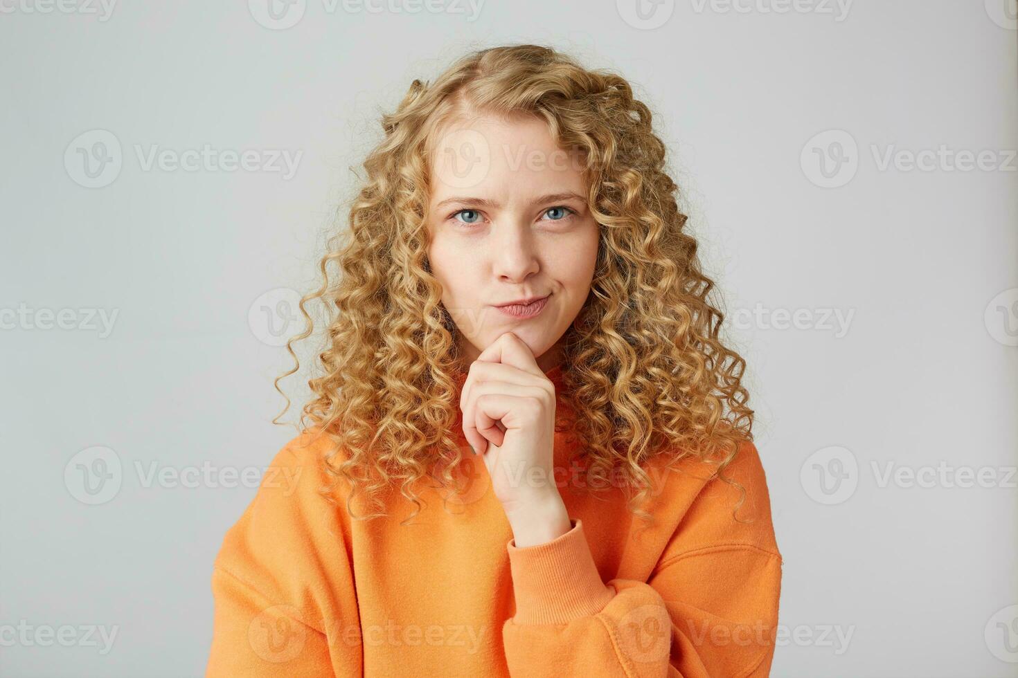 Horizontal shot of attractive young female with pensive contemplating expression,ponders about something,keeps hands in fist under chin,dressed in casual orange sweater, isolated over white background photo