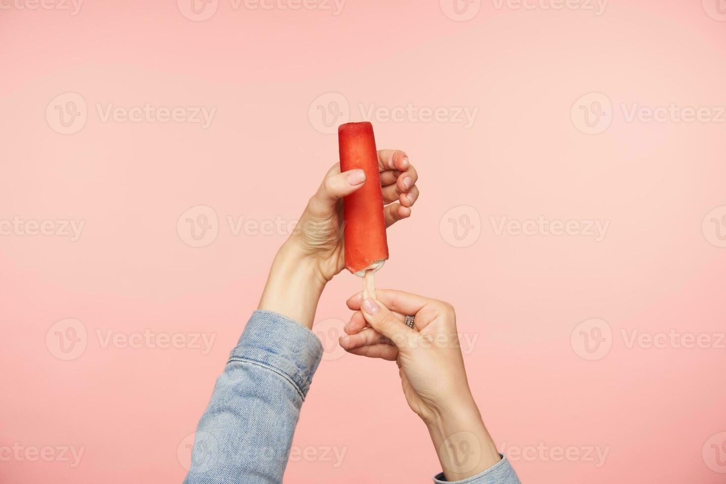 Horizontal shot of young female's hands with nude manicure posing over pink background with ice-cream on stick with red glaze. Food photography concept photo