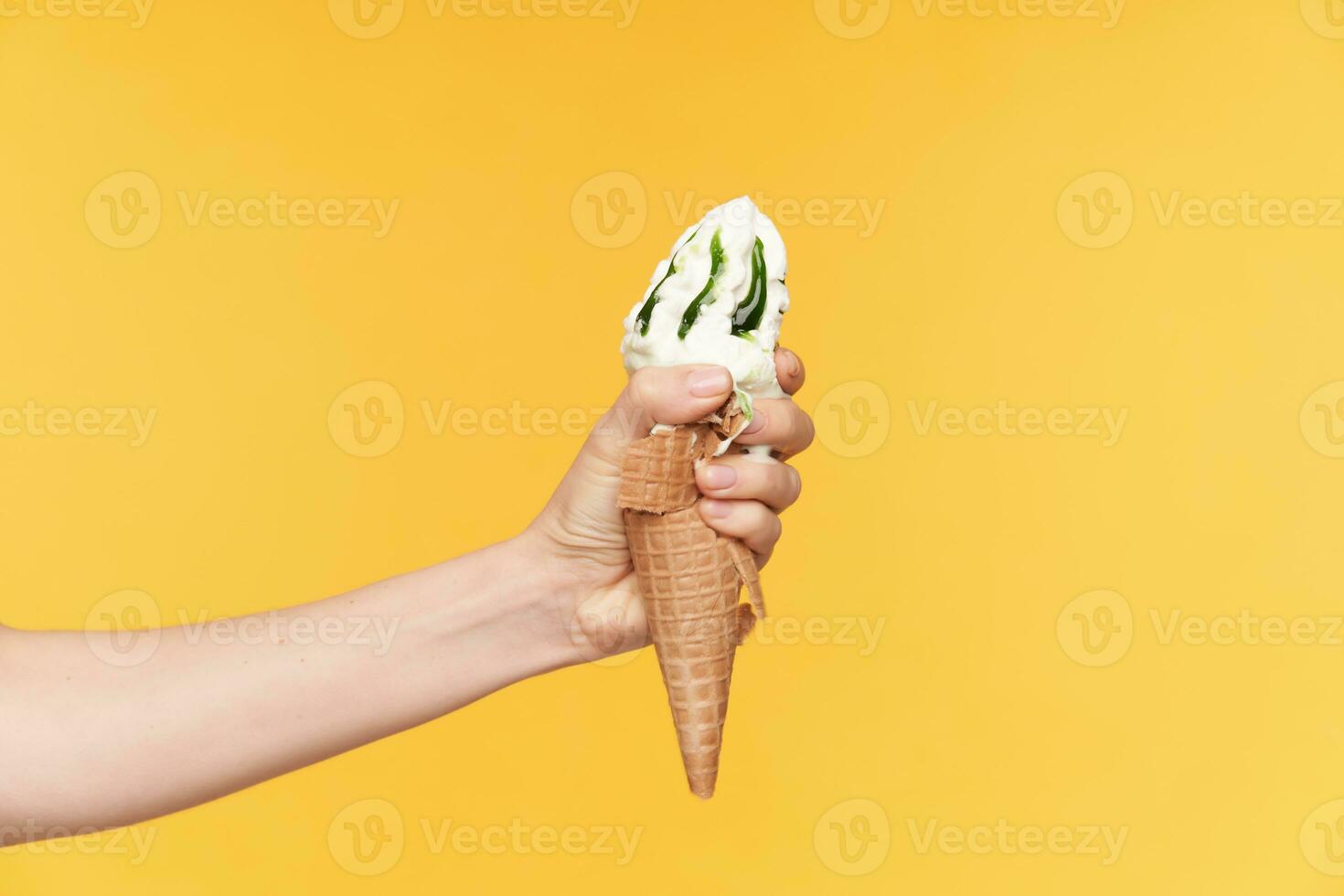 Horizontal shot of pretty fair-skinned female's hand smashing ice-cream while posing against yellow background. Food photography concept photo