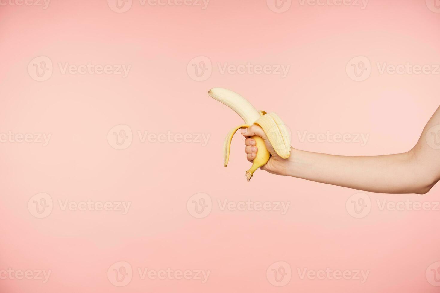 Side view of raised well-groomed female's hand with nude manicure holding peeled banana while posing over pink background, going to have healthy snack photo