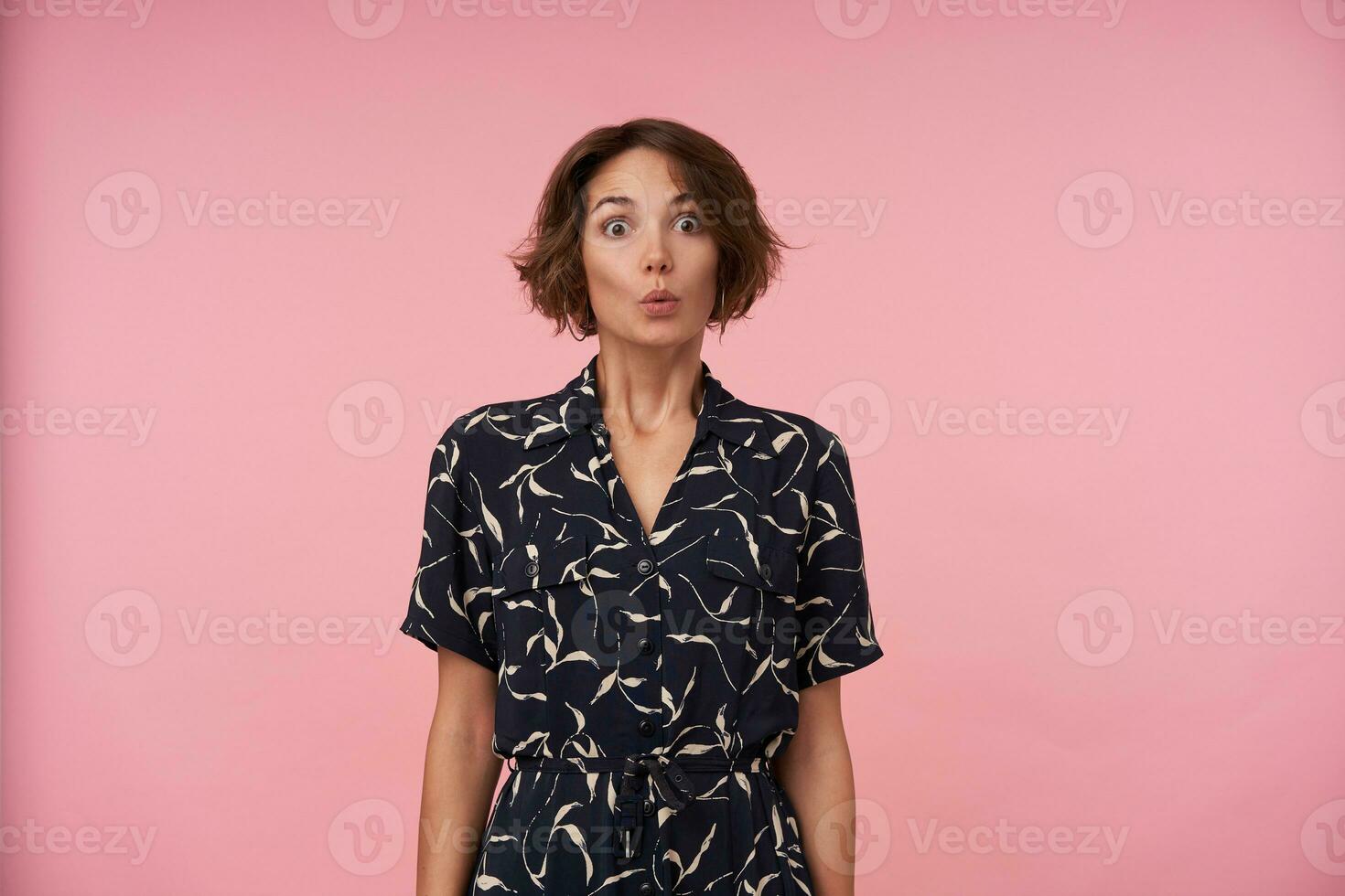 Portrait of pretty brunette lady in black patterned dress with short haircut posing over pink background with hands down, rounding eyes and raising eyebrows amazedly photo