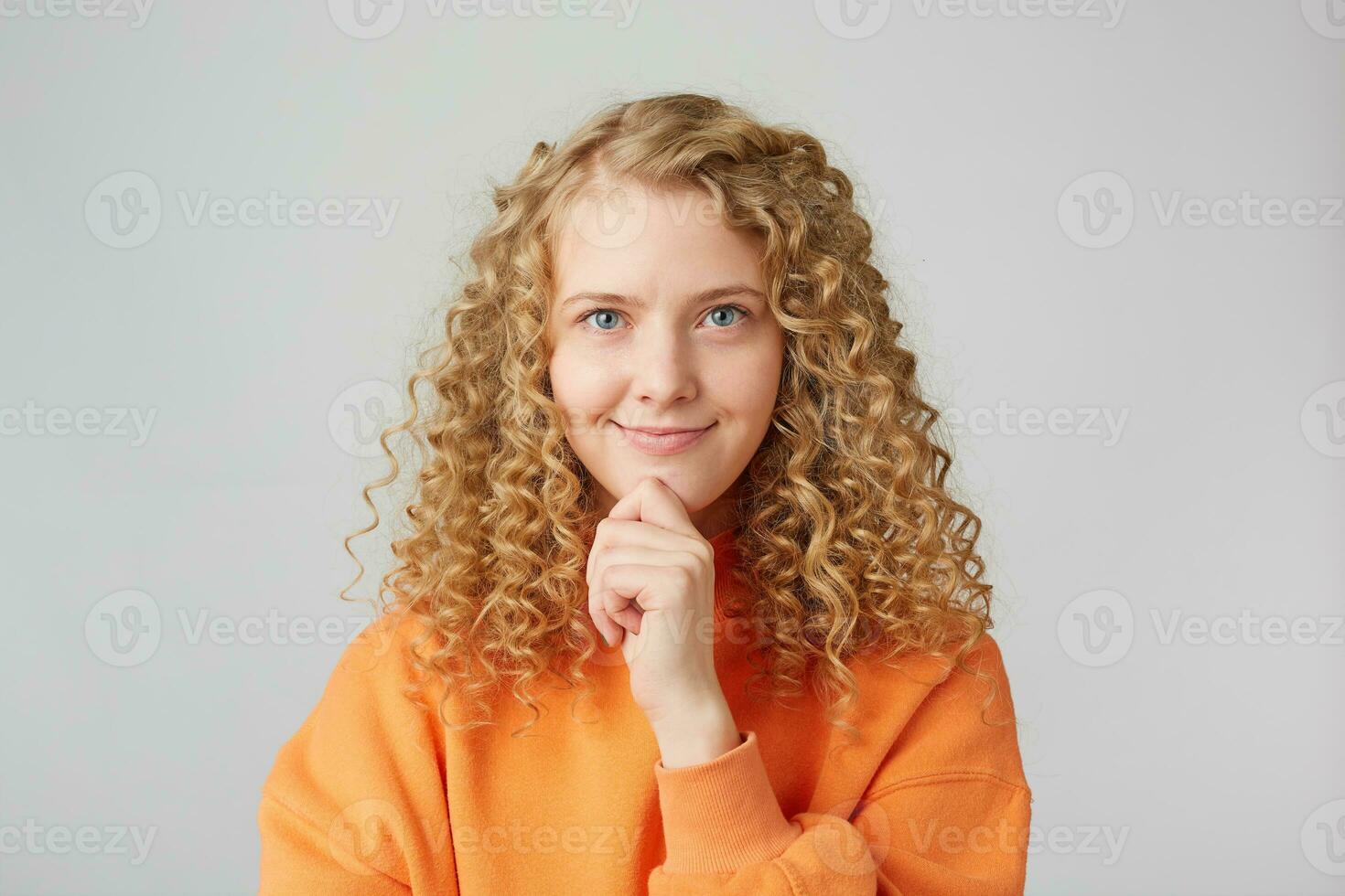 Close up shot of beautiful female model keeps hands in fist under chin, smiles, being in good mood after stroll or date with boyfriend, dressed in casual orange sweater, stands against white wall photo
