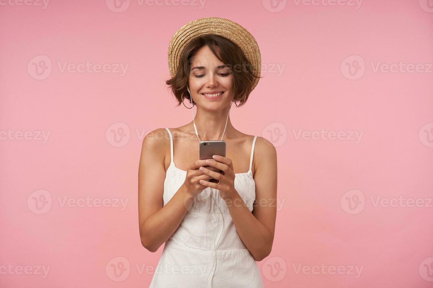 Studio shot of lovely young woman with short brown hair holding mobile phone in her hands and looking cheerfully on screen, watching pleasant videos with earphones, isolated over pink background photo