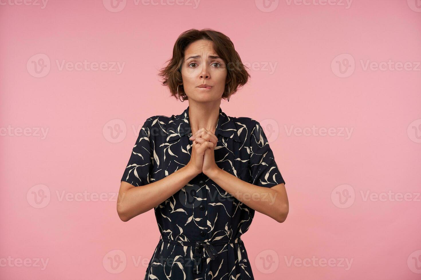 Studio shot of pretty brunette female with casual hairstyle wearing black patterned dress posing over pink background, looking at camera with desperate face and keeping palms folded photo