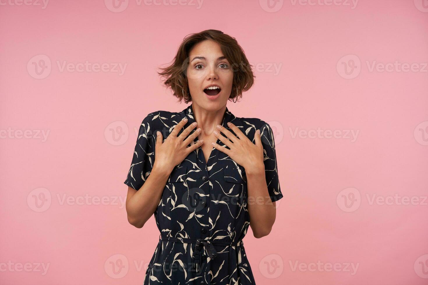 Surprised pretty brunette female with casual hairstyle posing over pink background and keeping palms on her chest, looking at camera with wide mouth opened, wearing black patterned dress photo