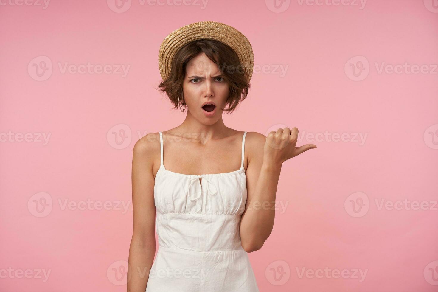 Portrait of young brunette female in white summer looking at camera with confused face, showing aside with thumb while standing over pink background, frowning her eyebrows with opened mouth photo