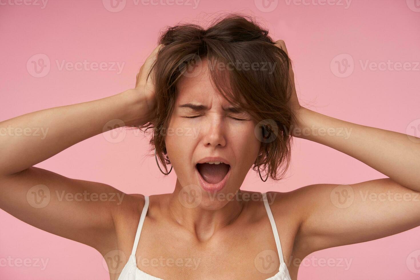 Stressed young woman with short brown hair covering ears with palms and closing eyes, frowning her face with wide mouth opened, wearing white top while standing over pink background photo