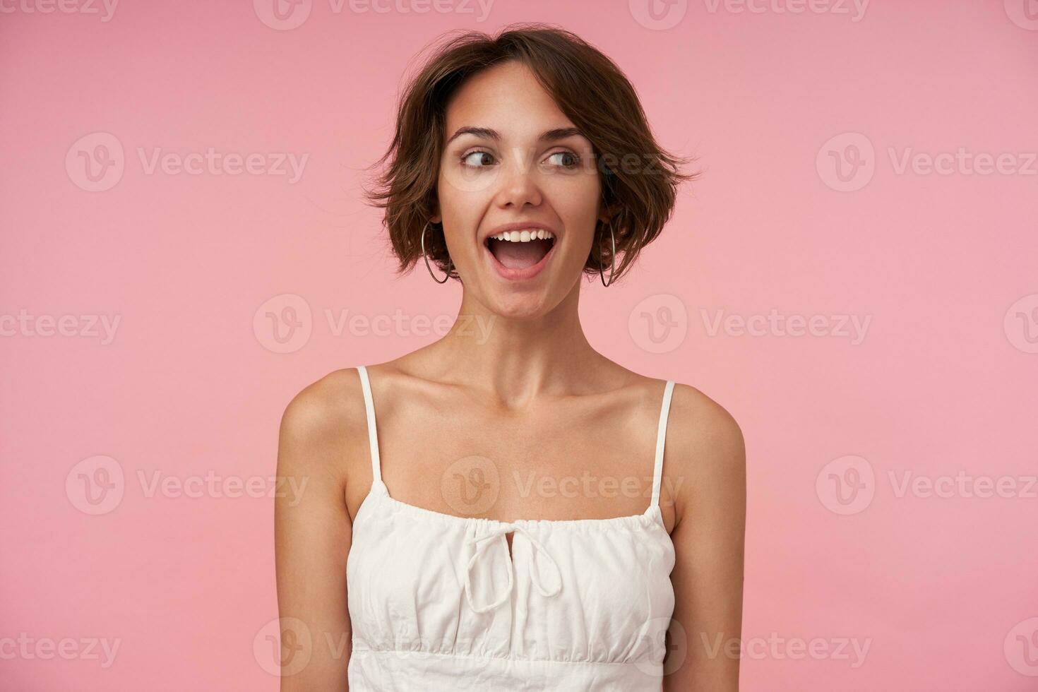 Joyful attractive brunette female with short haircut looking aside with wide happy smile, keeping hands along her body, posing over pink background, being in high spirit photo
