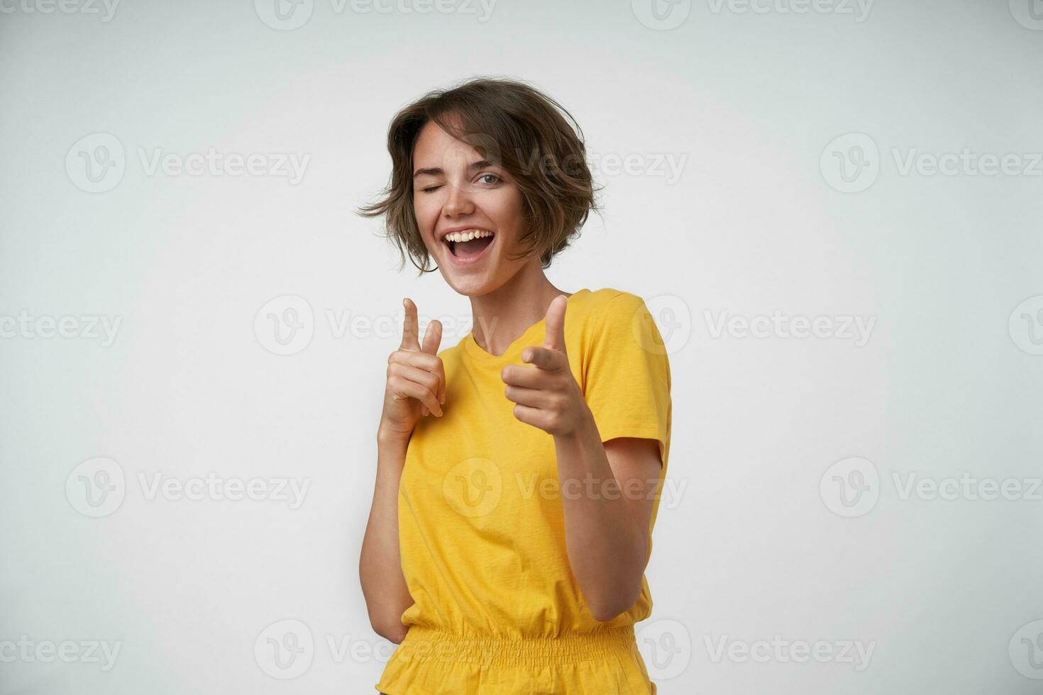 Positive young brunette woman with short haircut showing up her forefingers while posing over white background, giving wink to camera and smiling happily photo