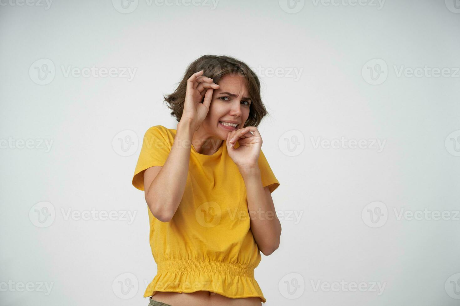 Indoor shot of afraid young female with short brown hair hiding behind raised hands, looking at camera scaredly and twisting her mouth, standing over white background in casual clothes photo