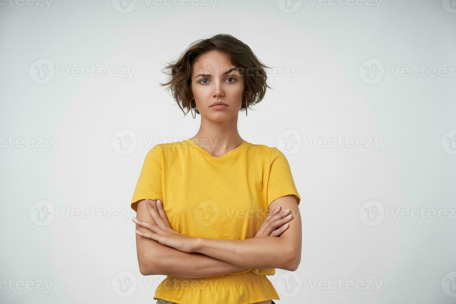 Indoor shot of young brunette lady with short haircut keeping her hands folded on chest, looking at camera and raising eyebrow confusedly, isolated over white background photo