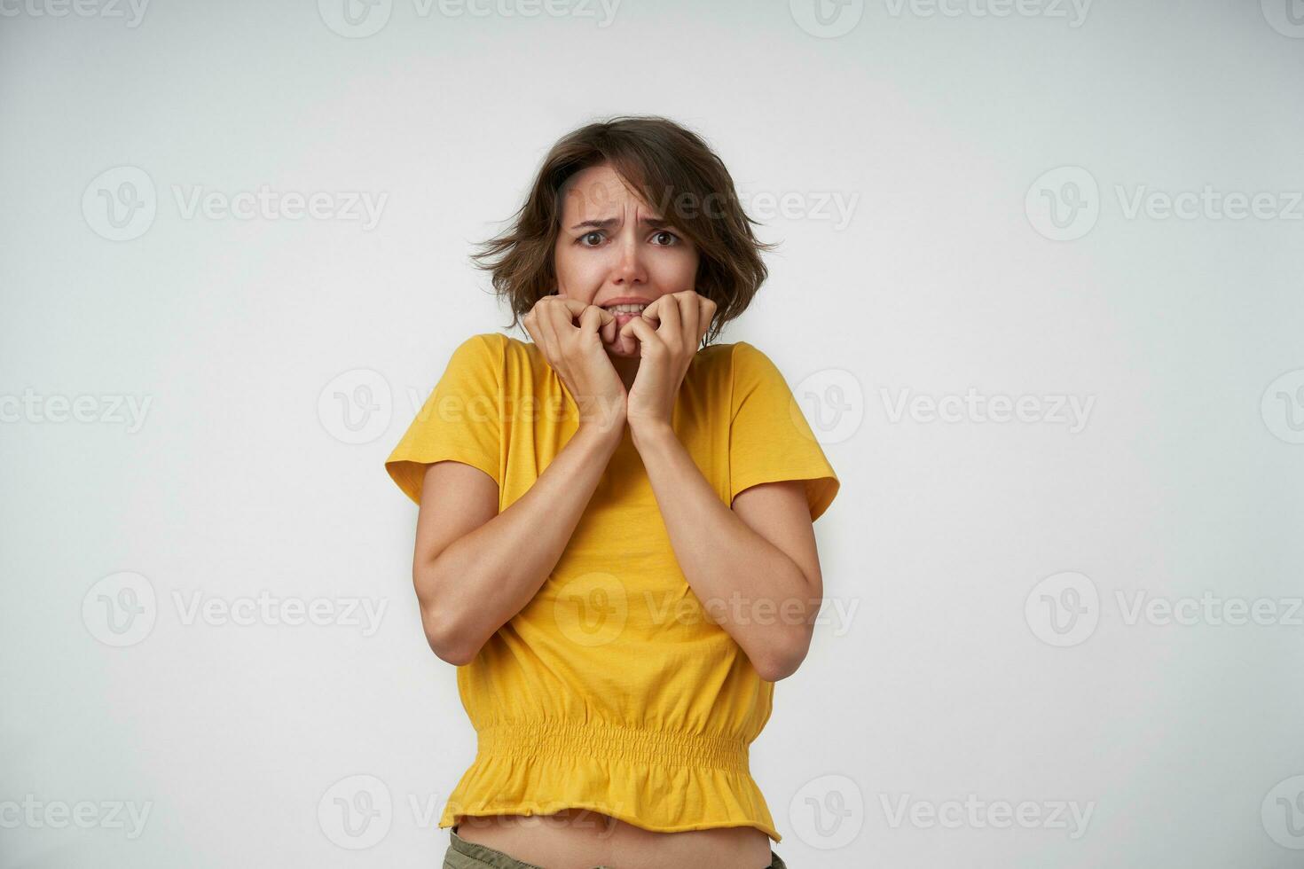 Studio shot of fearful young brunette female with short haircut keeping hands on her face while looking to camera scaredly, frowning face and showing teeth over white background photo