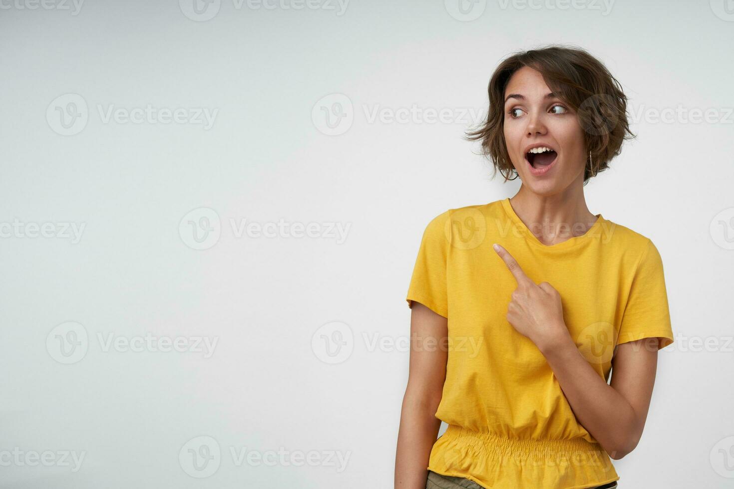 Surprised pretty brunette lady with short haircut looking aside with wide eyes and mouth opened, showing aside with forefinger while standing over white background photo