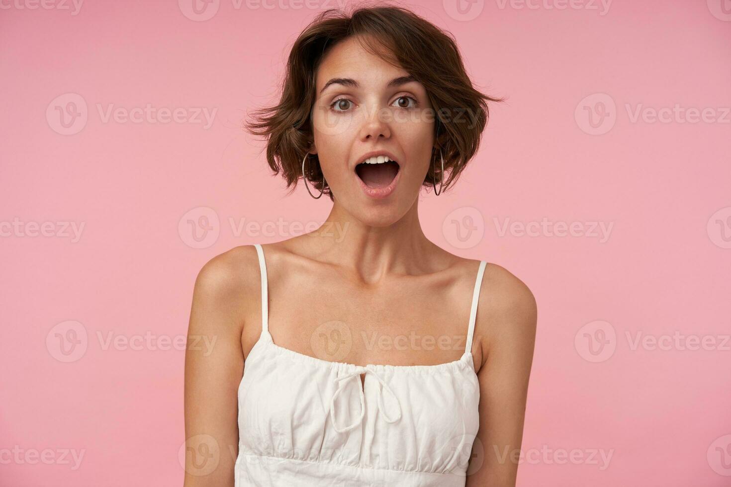 Surprised pretty young brunette woman with short haircut looking at camera with wide eyes and mouth opened, posing over pink background with hands down photo