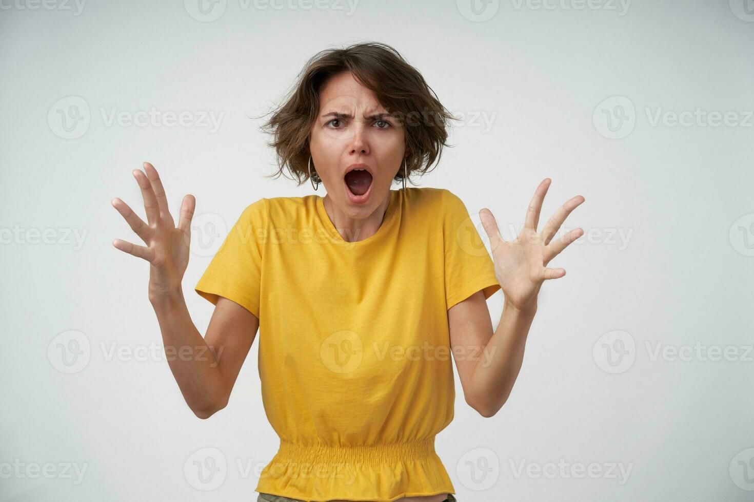Sressed brunette female with casual hairstyle looking at camera with wide mouth opened and raising palms scowlingly, frowning eyebrows while standing over white background photo