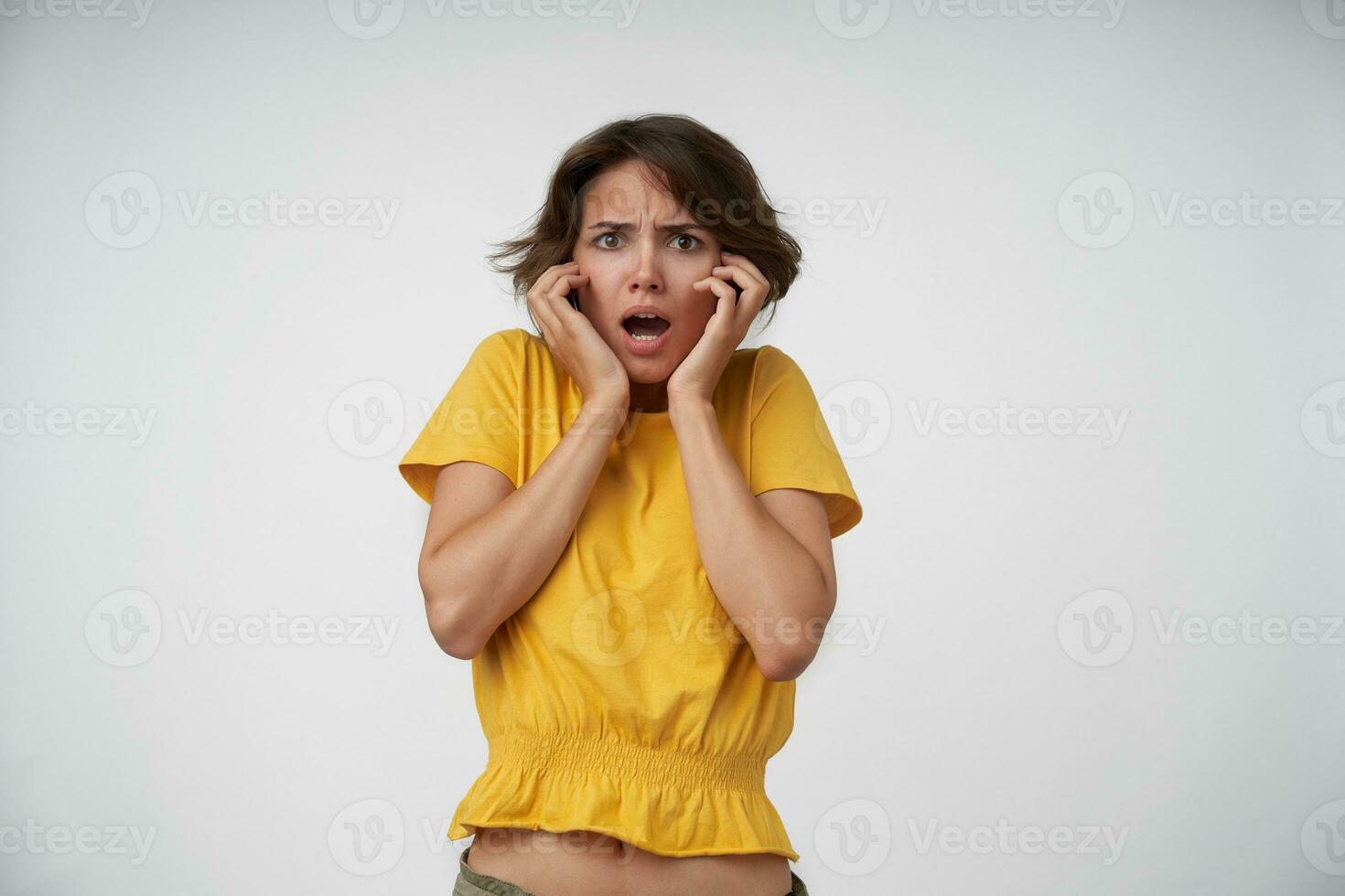 Funky young short haired female wearing yellow t-shirt while standing over white background, looking at camera with confused face, frowning eyebrows and keeping hands on her face photo