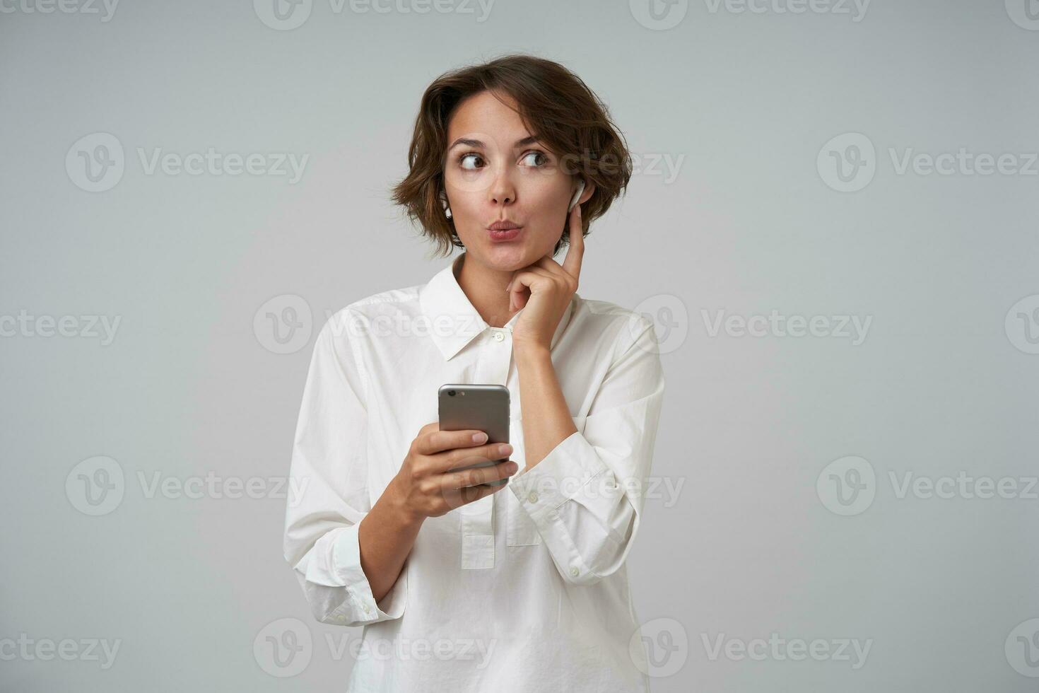 retrato de encantador joven morena hembra en blanco camisa escuchando a radio con su auriculares, acuerdo móvil teléfono en mano y mirando aparte con doblada labios, en pie terminado blanco antecedentes foto