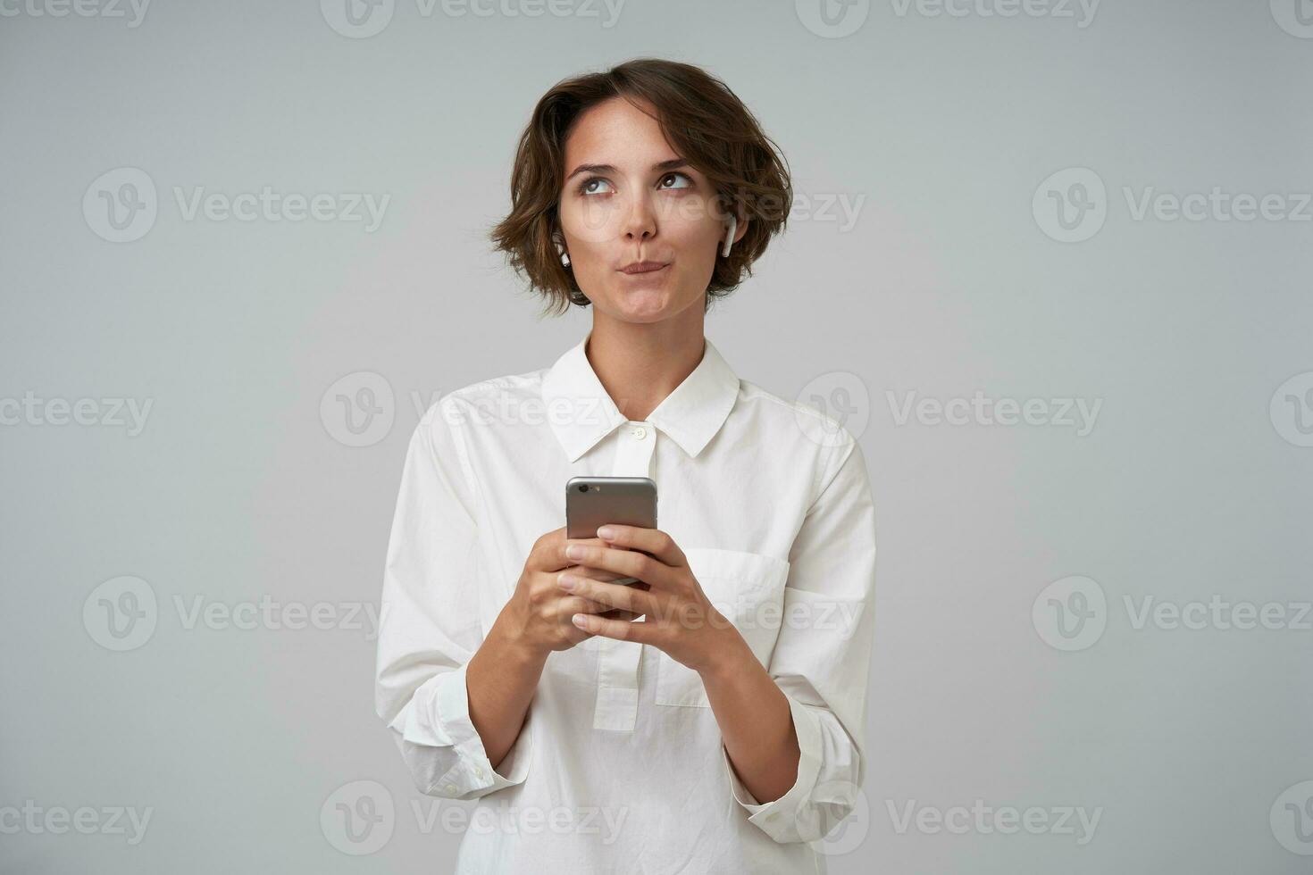 Horizontal shot of attractive young lady with short haircut wearing white shirt, looking aside dreamily while checking social networks with her smarthone, isolated over white background photo