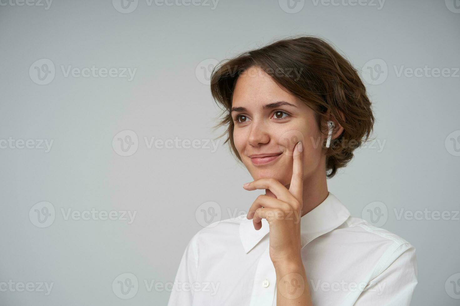 Positive young attractive woman with short haircut wearing white shirt while standing over white background, looking aside with charming smile and keeping forefinger on her cheek photo