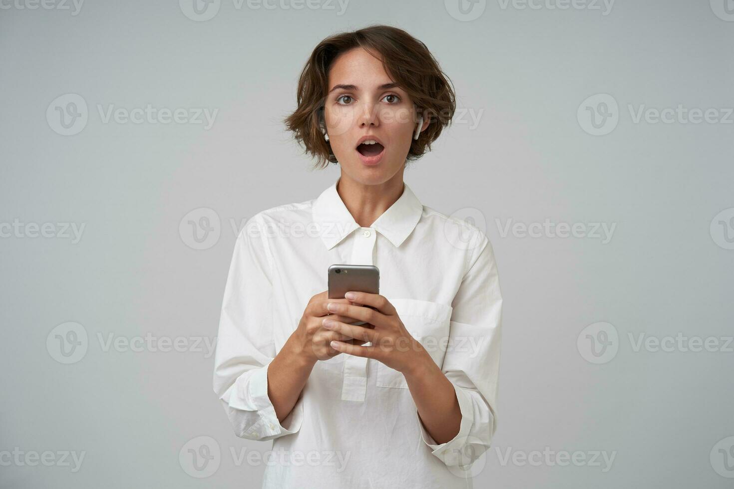 Portrait of surprised pretty brunette female with casual hairstyle holding mobile phone in hands while standing over white background, looking at camera with wide mouth opened photo