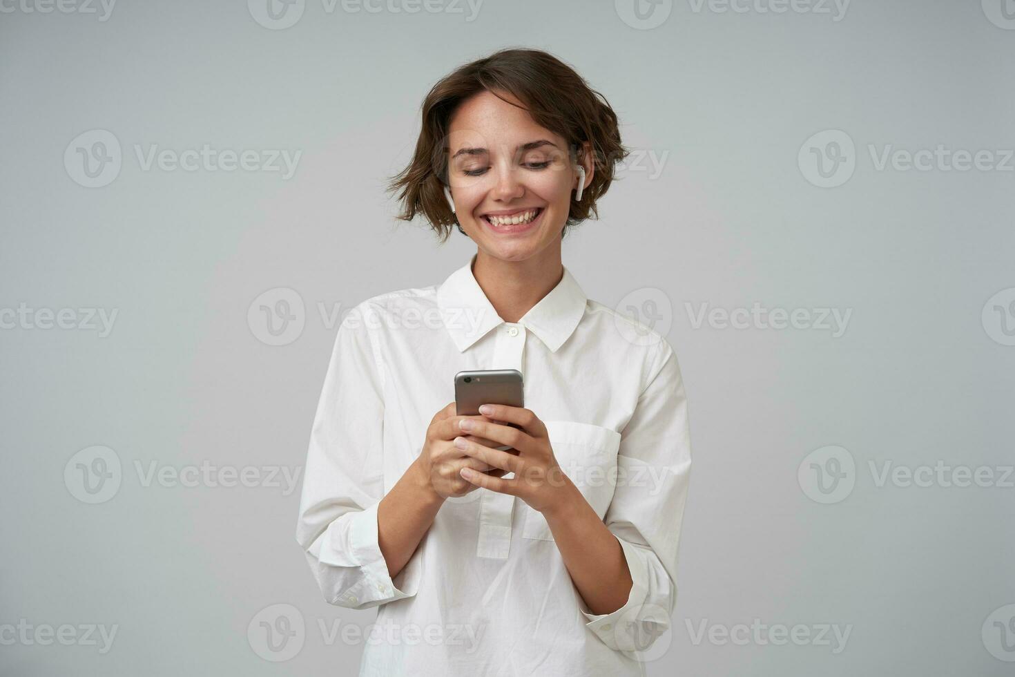 Cheerful young brunette lady with short haircut holding mobile phone in raised hands and looking happily at screen, wearing formal clothes while standing over white background photo