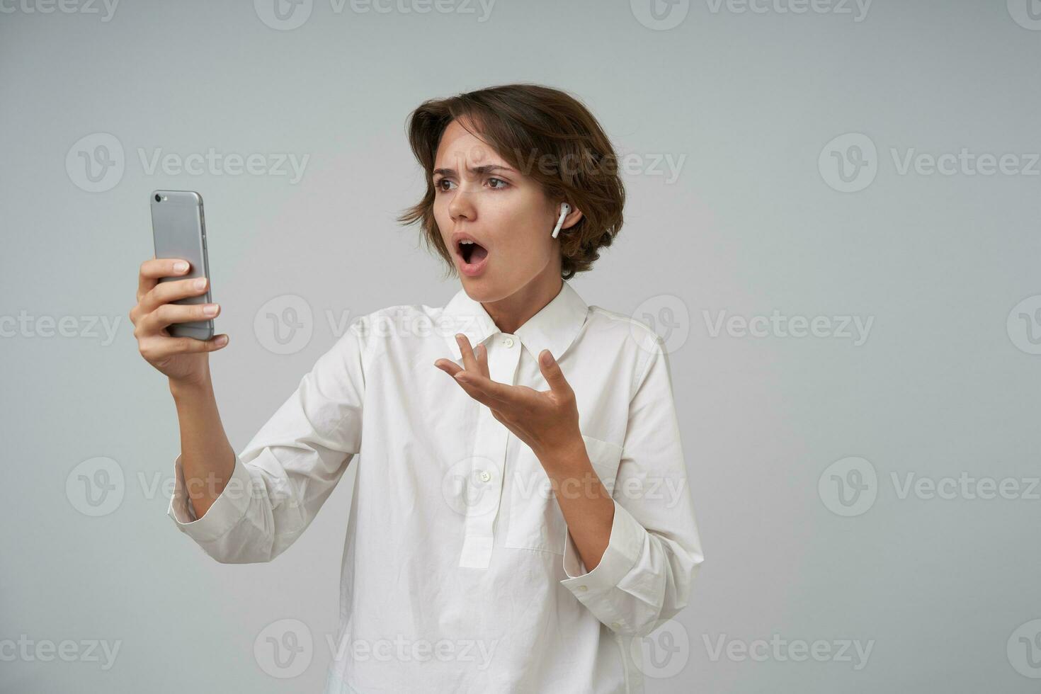 Portrait of young glum female with casual hairstyle holding mobile phone in raised hand and having video call, looking at screen with shocked face, posing over white background in formal clothes photo