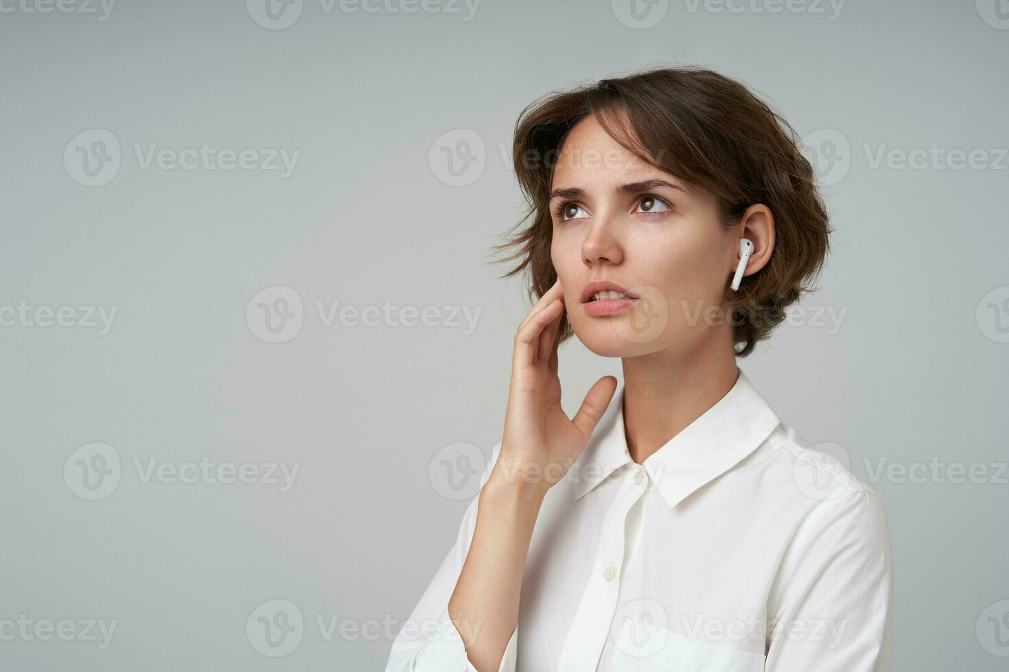 Portrait of serious young female with short haircut holding raised palm on her cheek and looking upwards thoughtfully, wearing white shirt while posing over white background photo