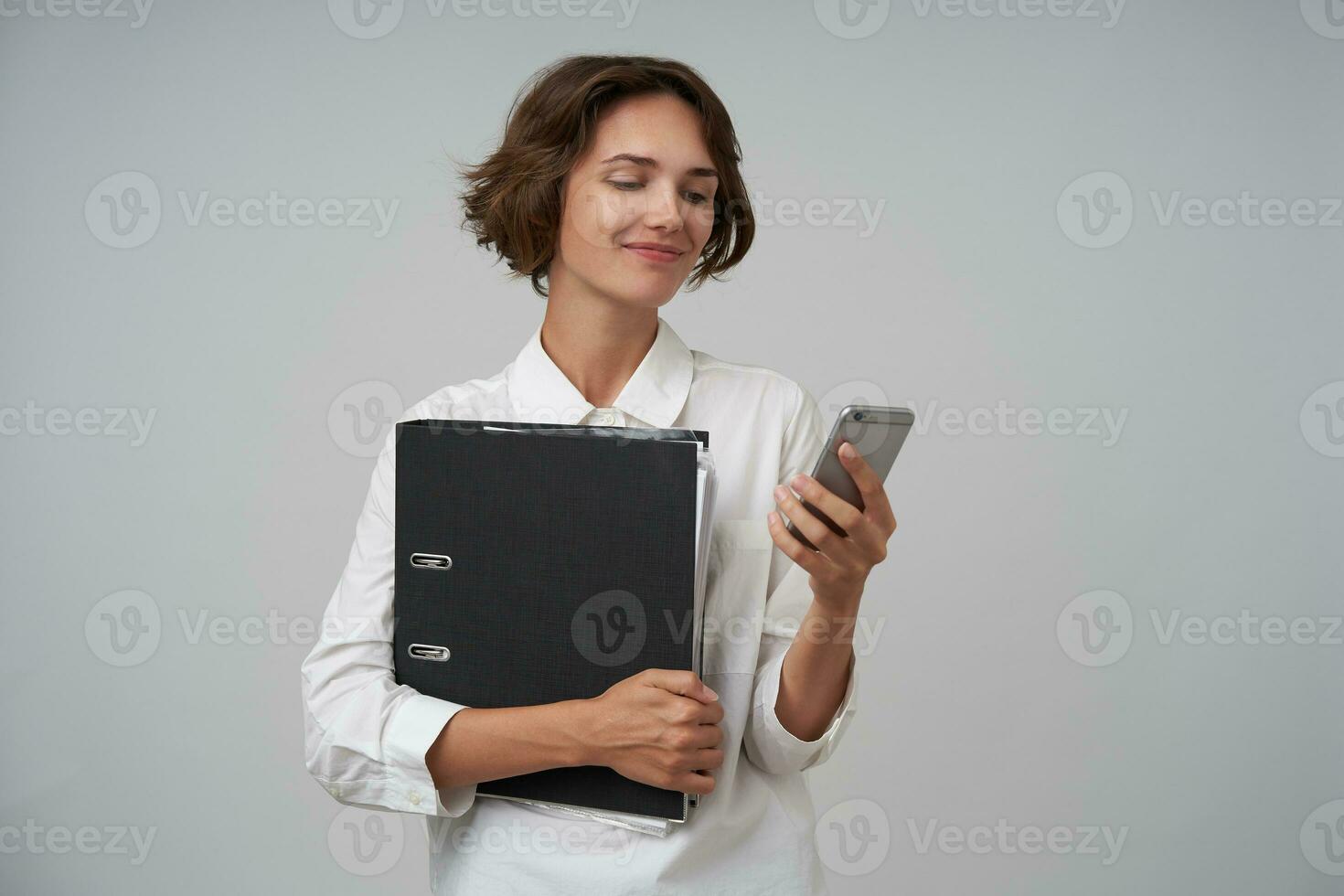 Portrait of pretty brunette woman with short haircut holding folder with documents and typing message on her mobile phone, being in nice mood, isolated over white background photo