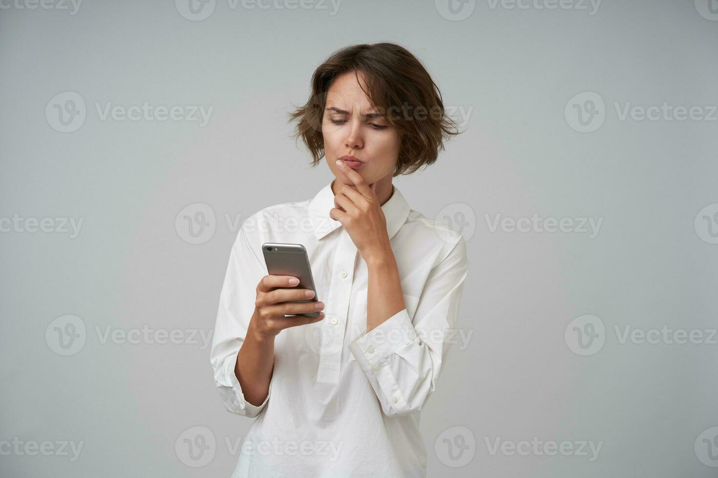 Severe young attractive woman with short haircut wearing formal clothes, keeping mobile phone in hand and looking seriously at screen, holding chin with hand while posing over white background photo
