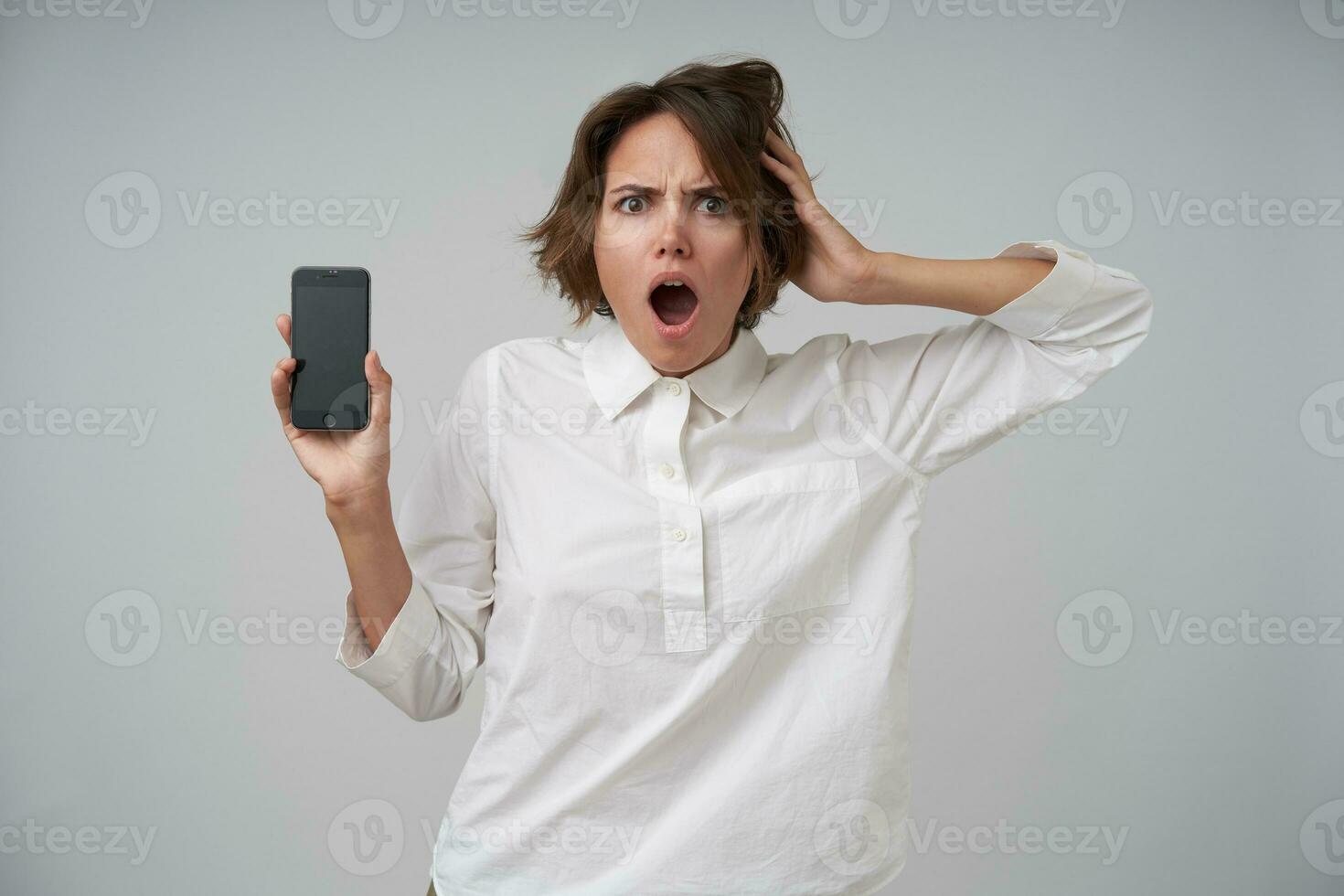 Shocked young brunette lady with casual hairstyle clutching her head and looking at camera surprisedly, holding mobile phone in raised hand while posing over white background photo