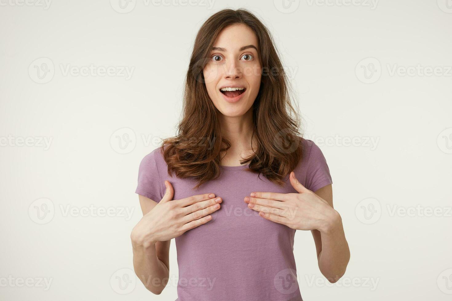 Portrait of charming young woman points at herself being puzzled to be chosen. Pretty attractive European female stands over white background, has confused expression. Surprisment concept photo