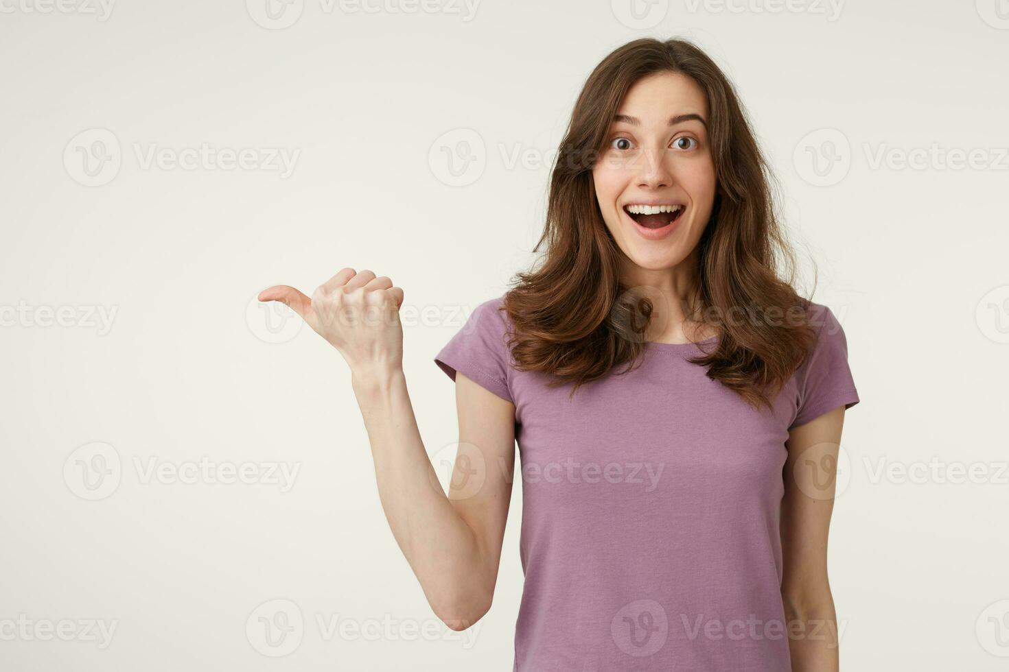 Beautiful young woman fascinated and excited. Close up of amazed attractive young female in t-shirt pointing aside with dropped jaw, standing over white wall, promotion and advertisement photo